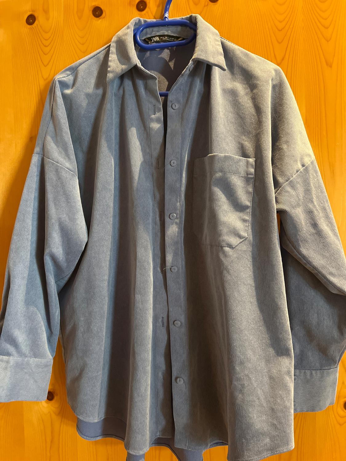 Light blue long-sleeved corduroy shirt with a pocket, hanging on a blue hanger against a wooden background. Label with the text ZARA.