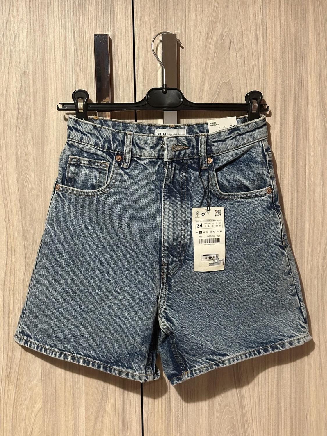 Blue denim shorts on a black hanger in front of a wooden wardrobe. Text on labels: ZARA, HI-RISE BERMUDA, 34, €19.95.