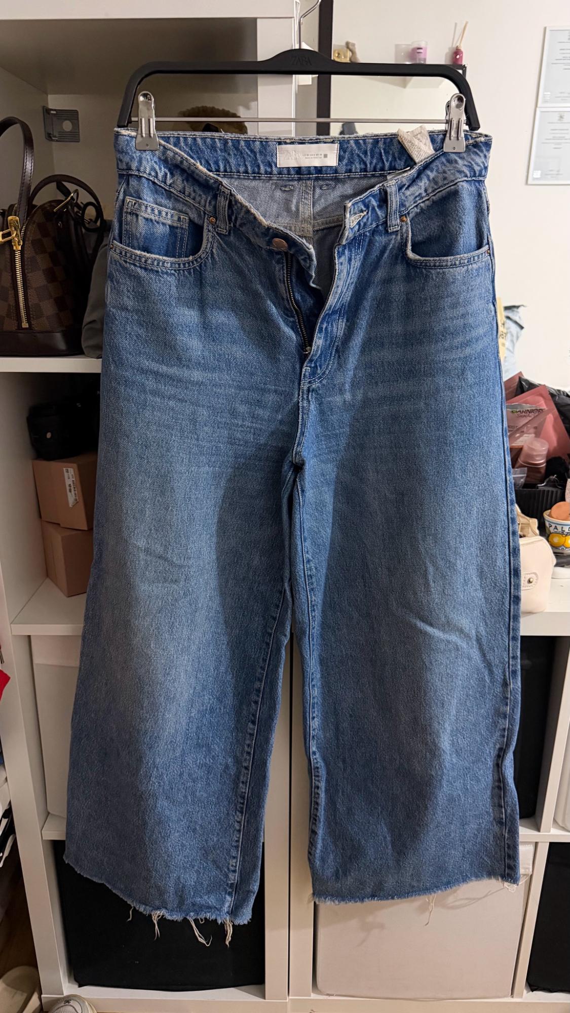 Wide-leg blue jeans with a double waistband and frayed hem, hanging on a hanger with the text ZARA in front of a white shelf.