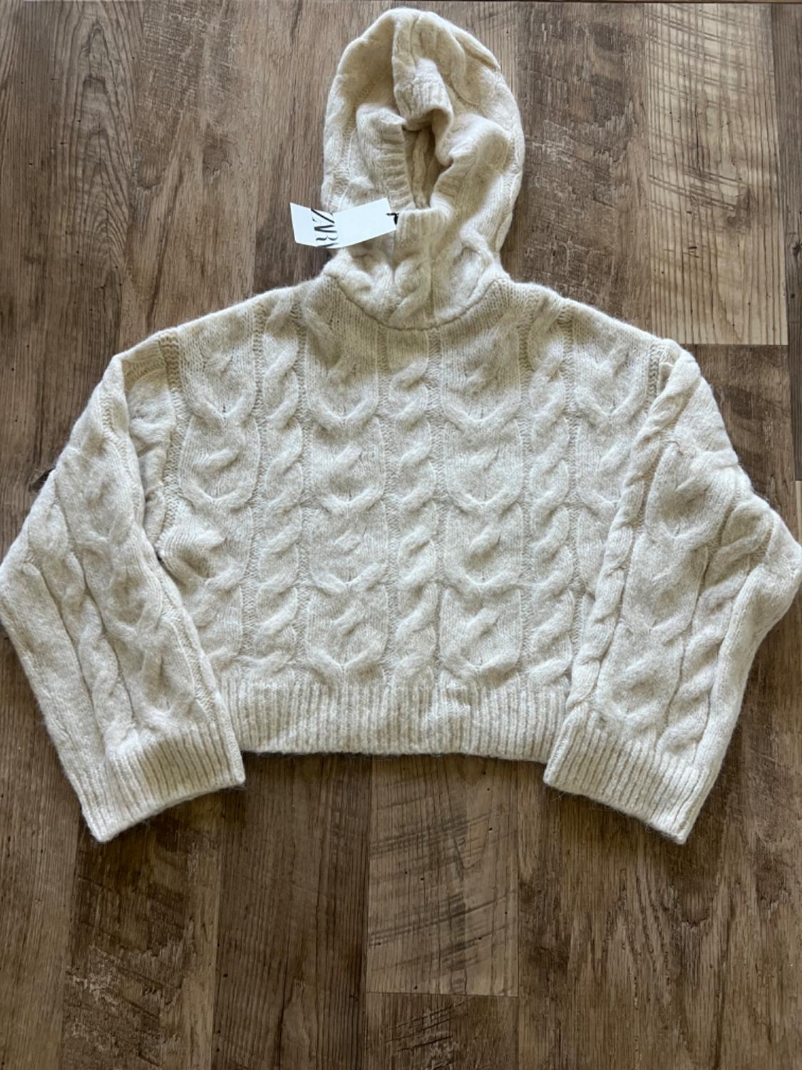Chunky cream knit hoodie, on a wooden floor. White label visible on the collar.