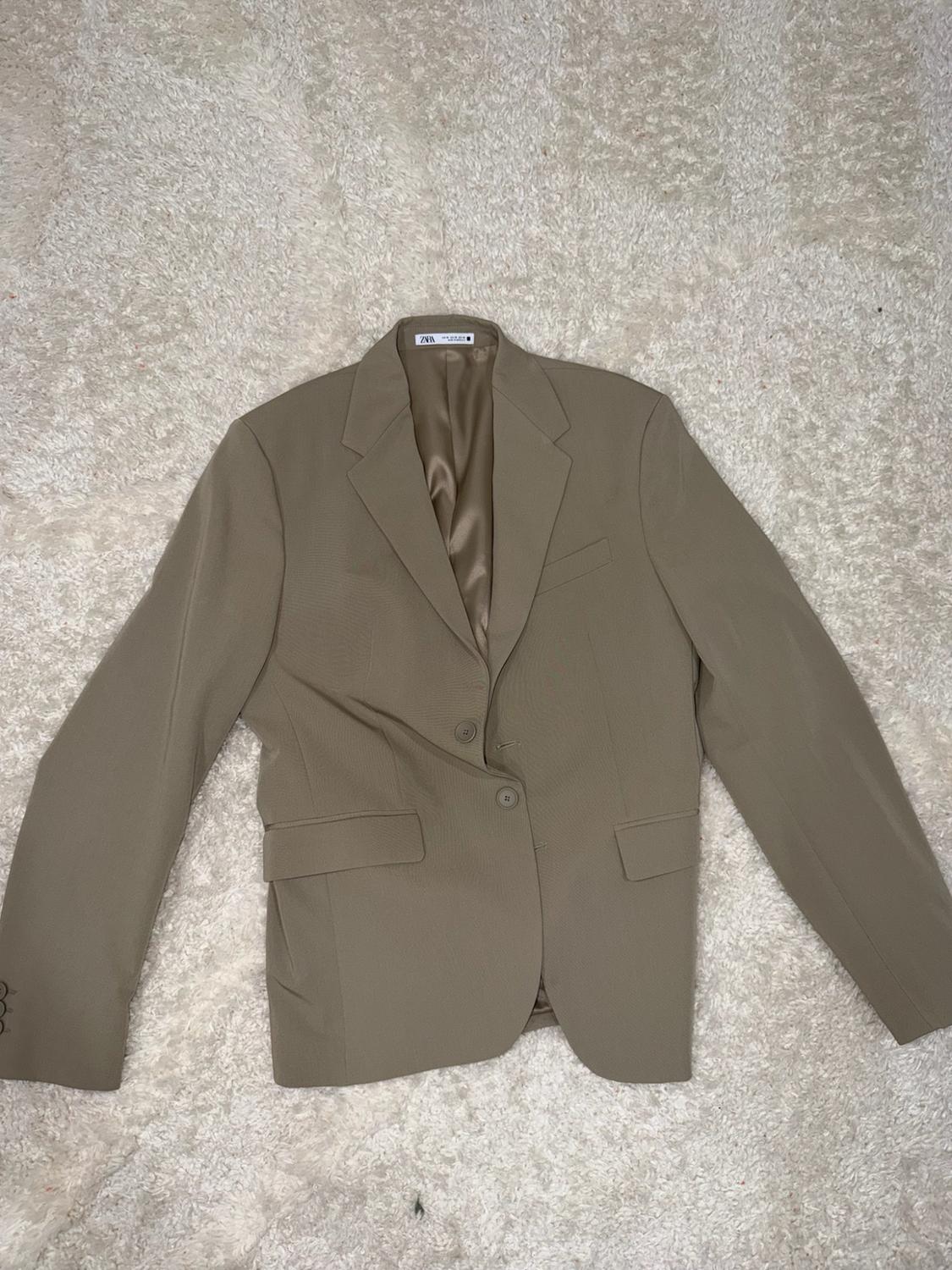 Beige two-button blazer with flap pockets, laid on a soft white rug. ZARA can be read on the label.