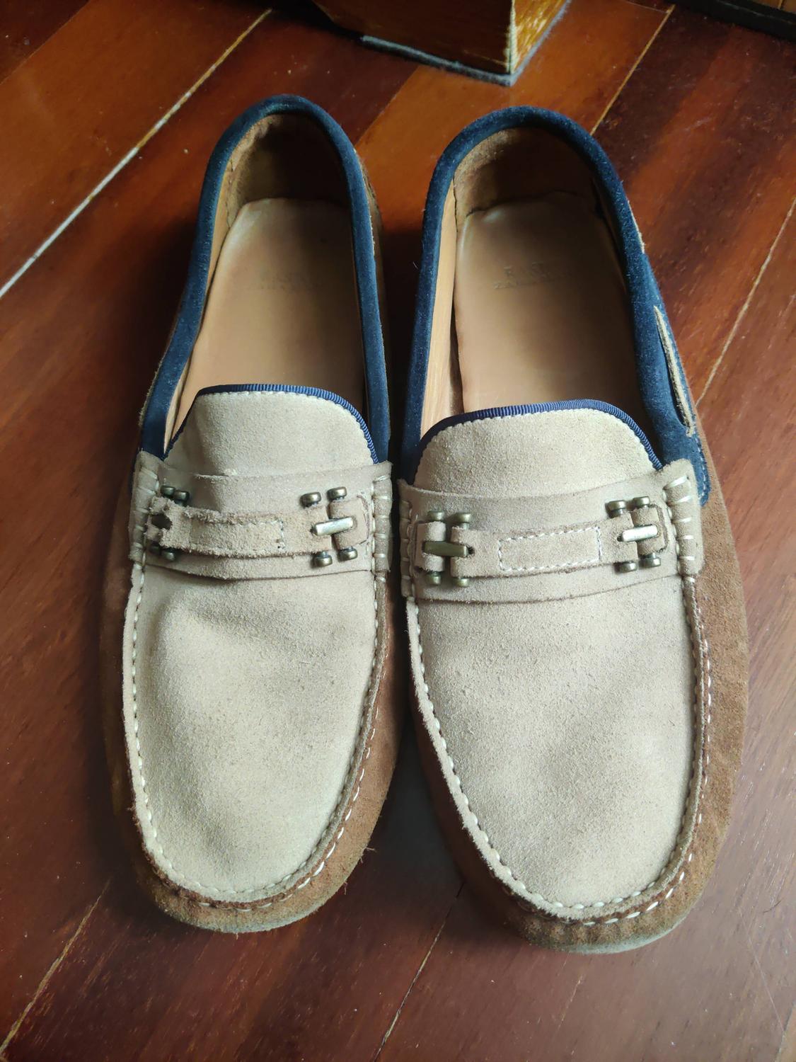 Beige and brown suede casual loafers with white stitching, blue trim and metallic appliqué, presented on a polished wooden floor.