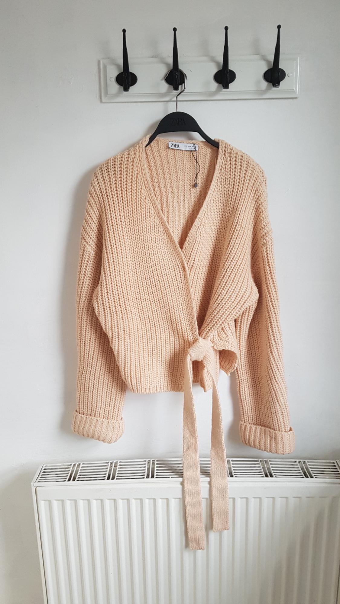 Beige knitted cardigan hanging on a black hanger with a visible label, over a white radiator and white wall background.