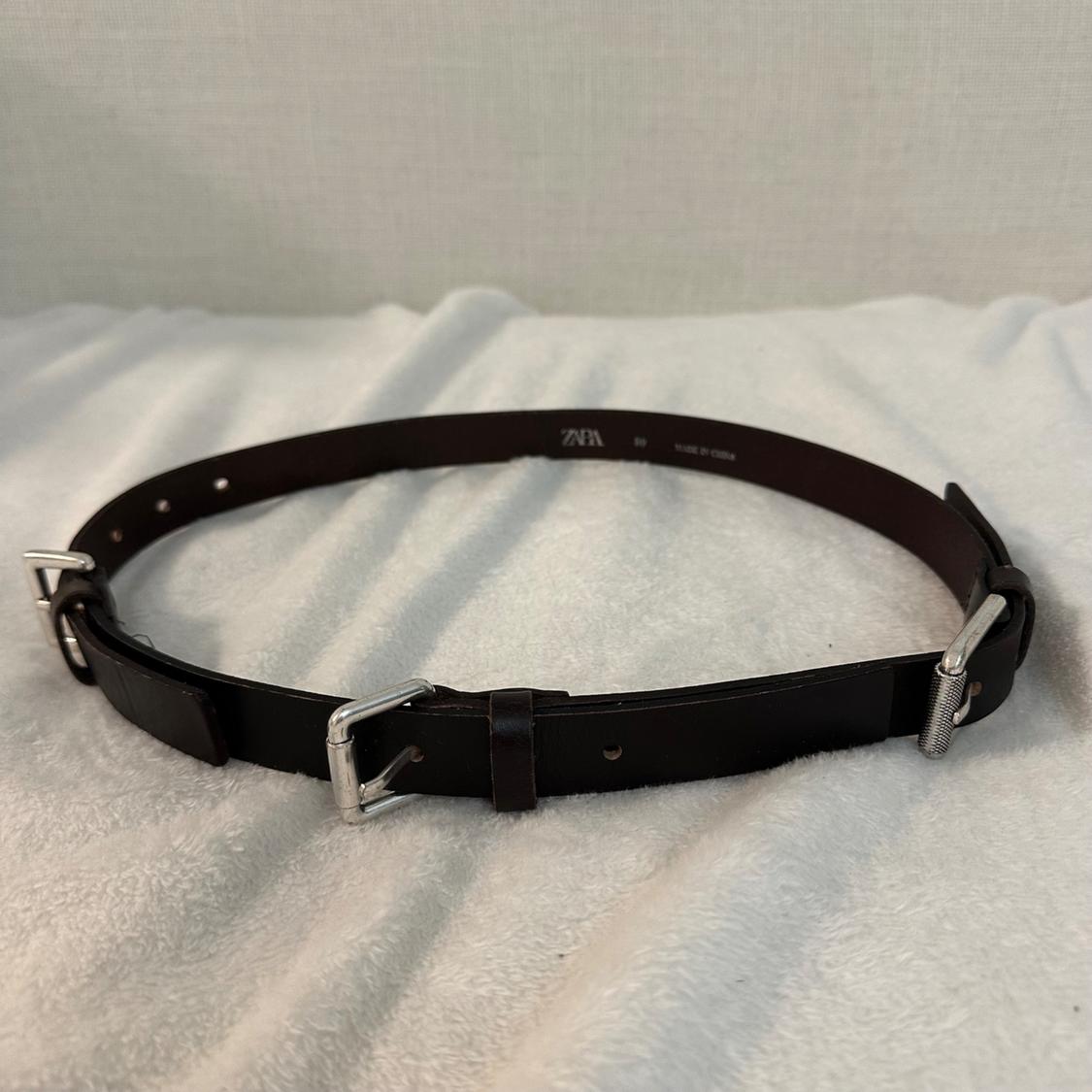 Black leather belt with silver buckles on a white surface. Visible text: "ZARA".