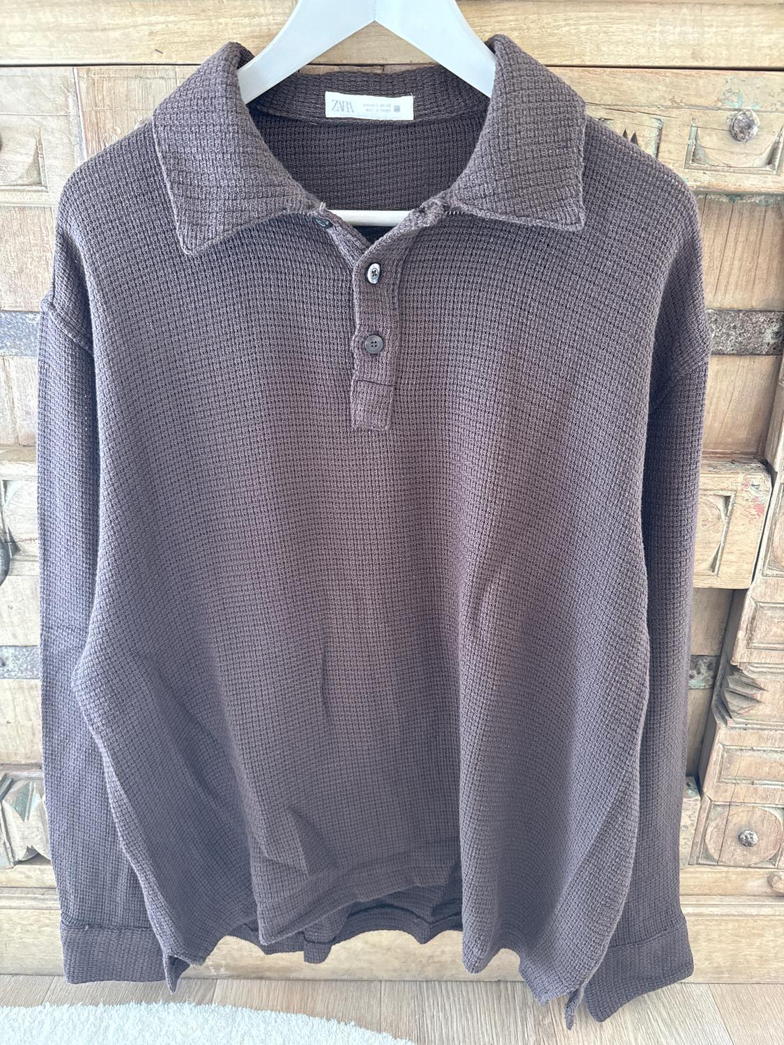 Brown textured fabric shirt with collar and buttons, hanging on a white hanger in front of a wooden cabinet.