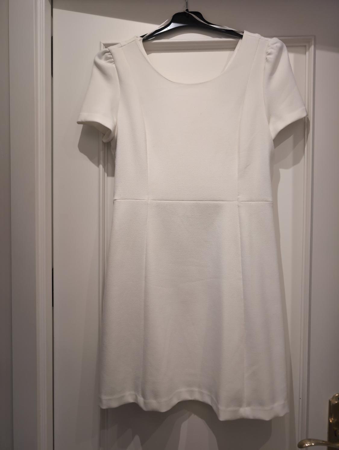 White, short-sleeved, round-neck dress hanging on a black hanger over a white panelled door with a gold doorknob.