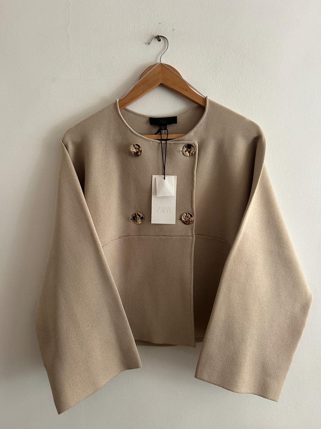 Beige long-sleeved jacket with decorative buttons, hanging on a wooden hanger. Visible label with the text "ZARA".