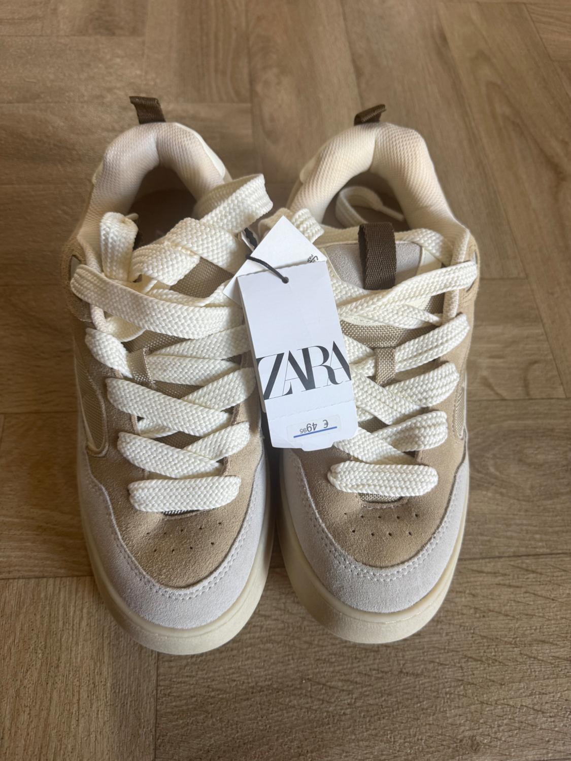 Beige and white trainers with Zara price tag on a wooden floor.
