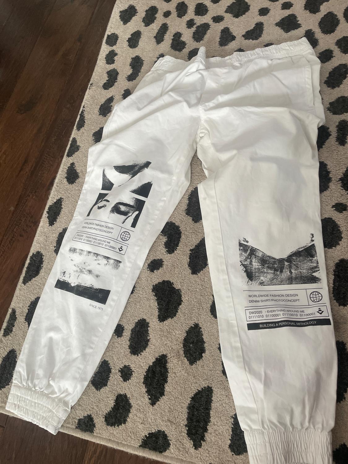 White joggers with black prints and the text WORLDWIDE FASHION DESIGN DENIM SHIRT/PHOTOCONCEPT on a speckled carpet.