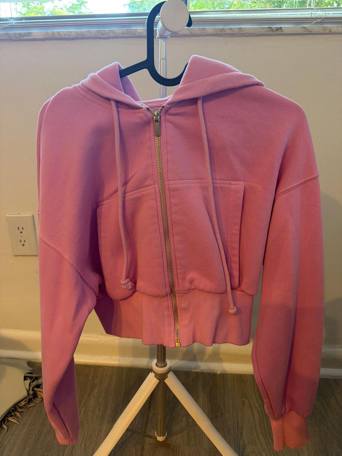 Pink cropped hoodie with pockets and a gold zipper, hanging on a hanger on a tripod in a casual interior.
