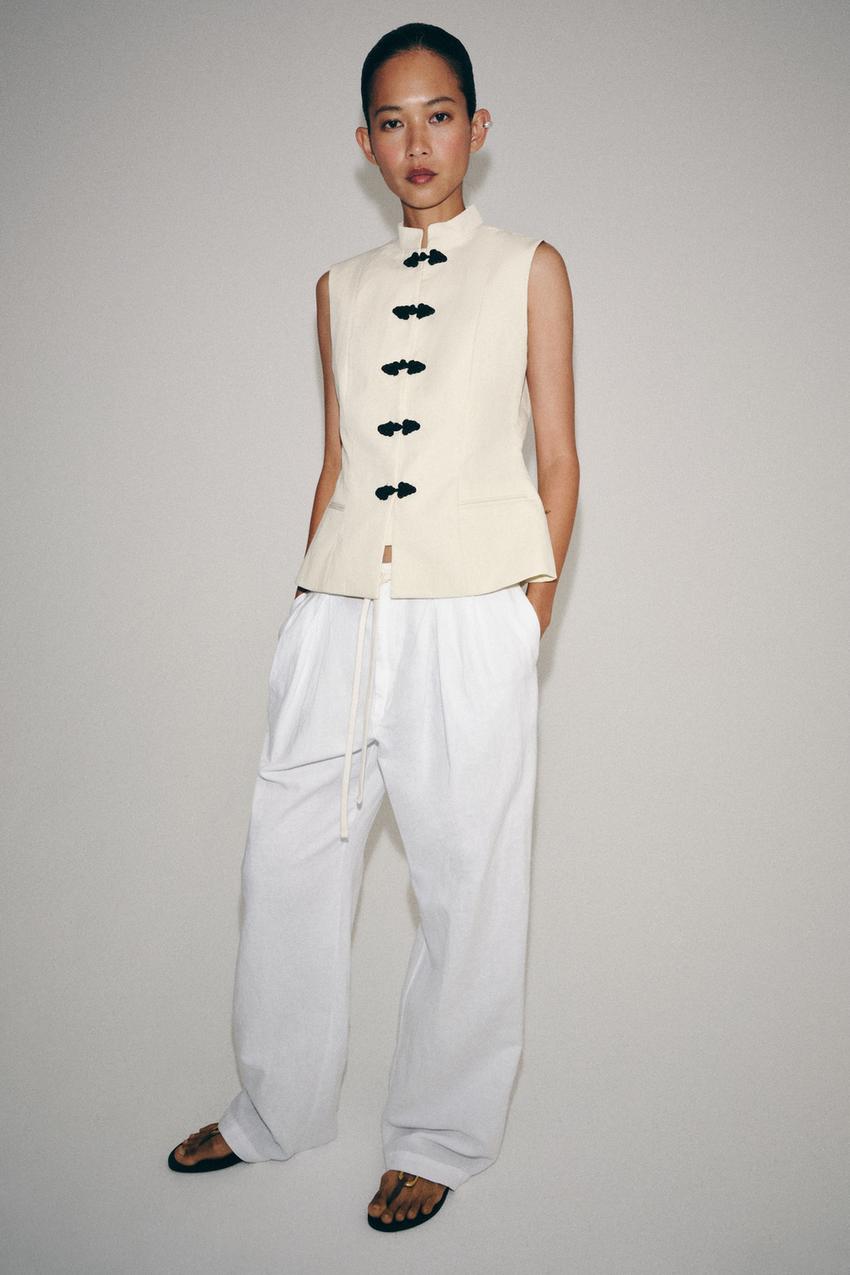 Cream sleeveless vest set with a stand-up collar, black frog closures, and front pockets, paired with wide white pants and a drawstring.