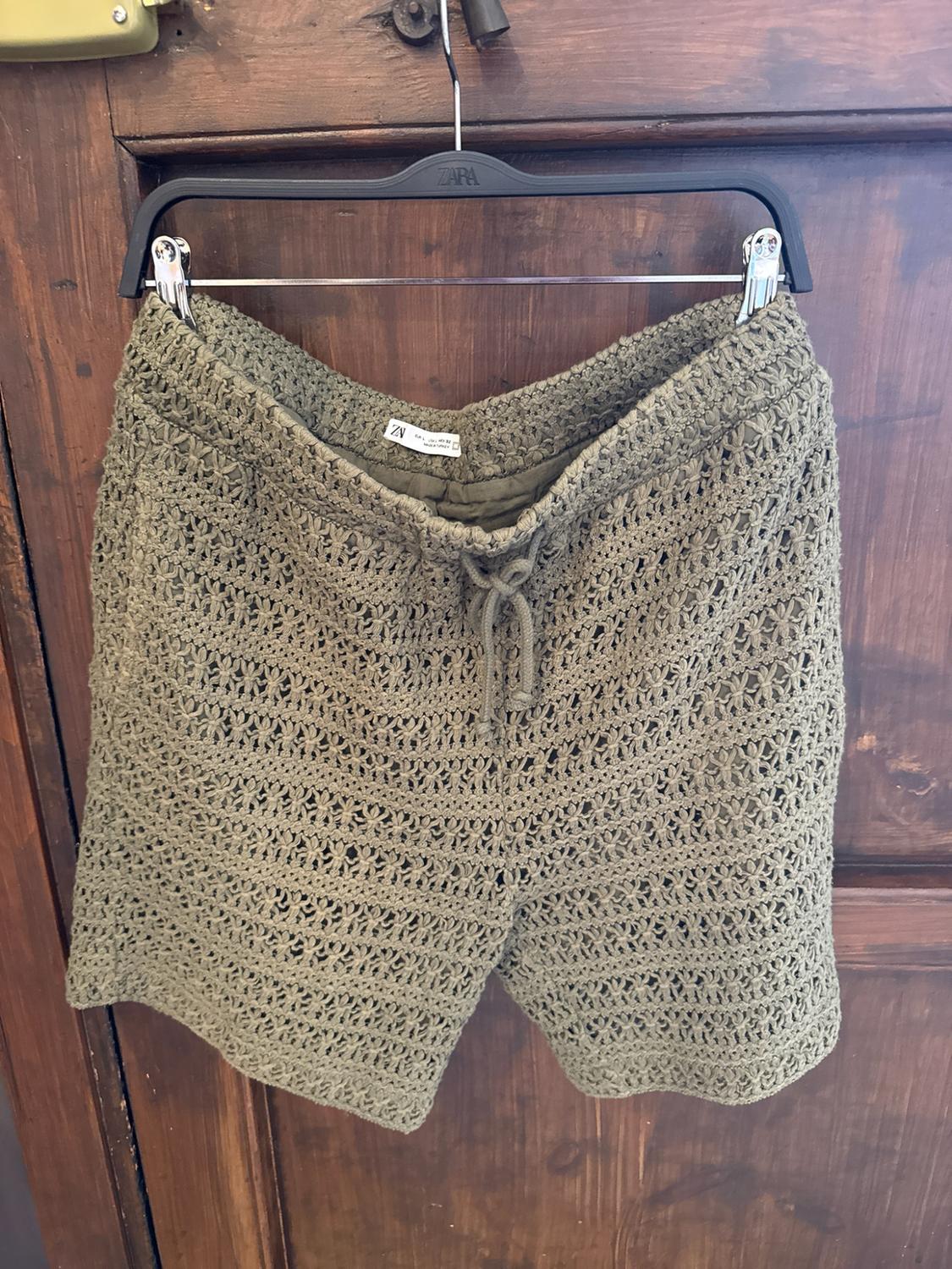 Green open-knit shorts with a drawstring, hanging on a black hanger on a dark wooden door. Text: ZARA.