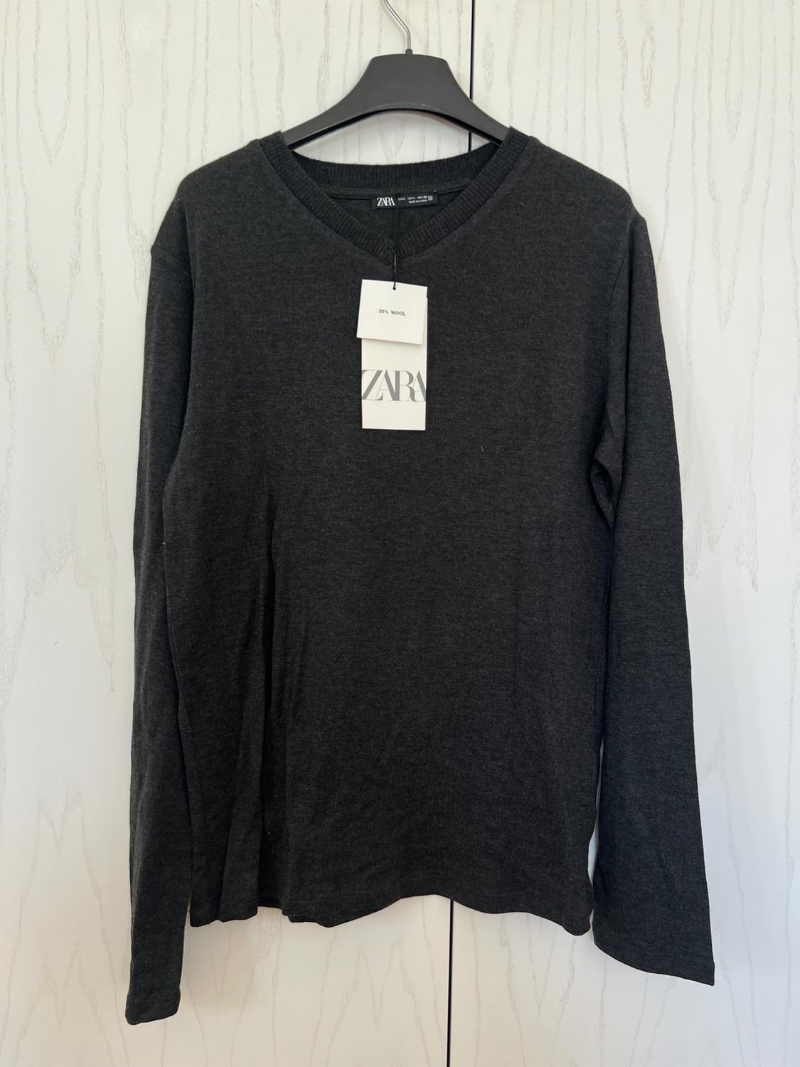 Dark grey, long-sleeved jumper hanging on a black hanger. Visible label with text "20% WOOL" and "ZARA". White wall background.