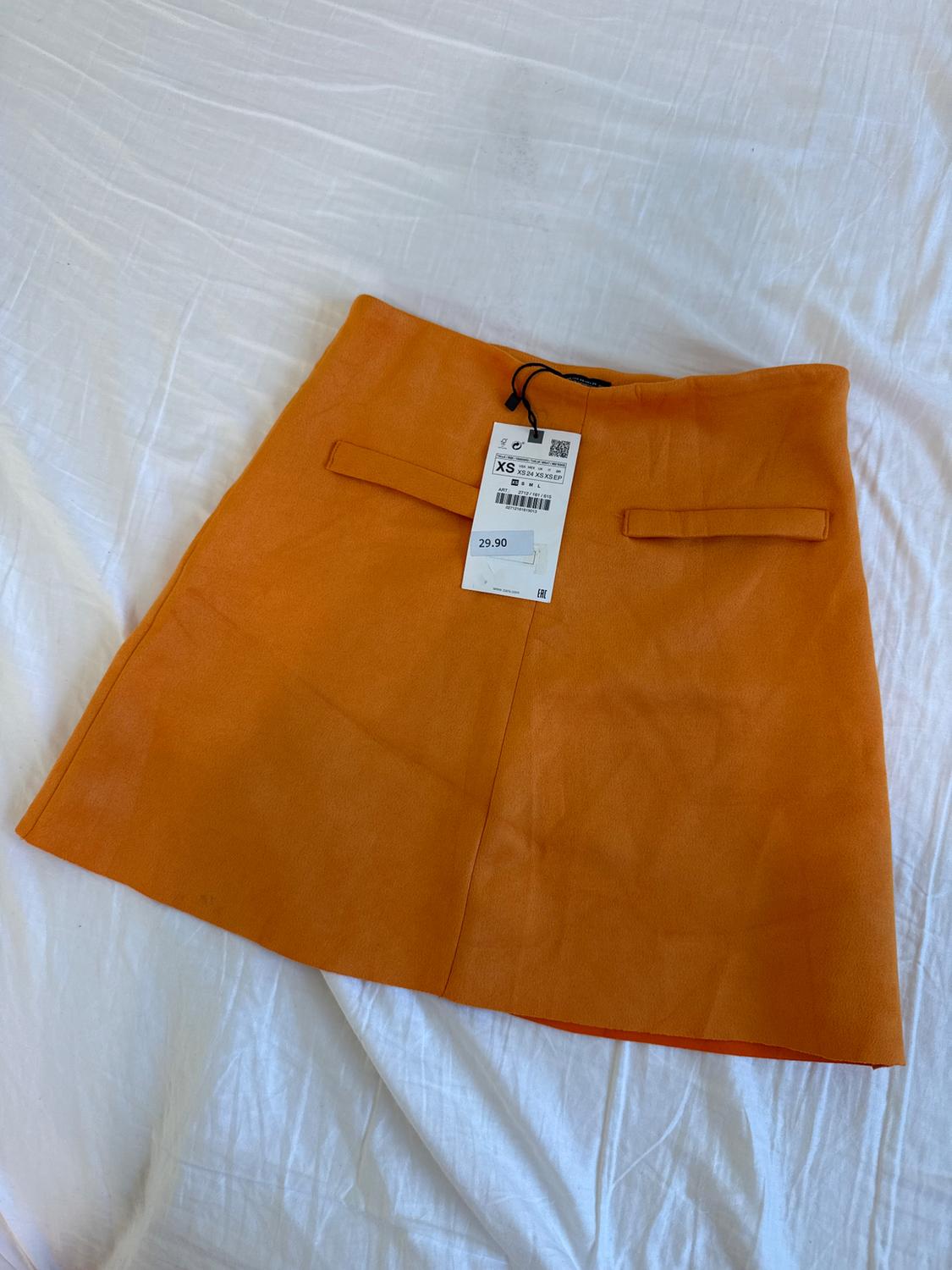 Orange skirt on white fabric, with a price tag of 29.90 and size XS. The skirt has two decorative back pockets.