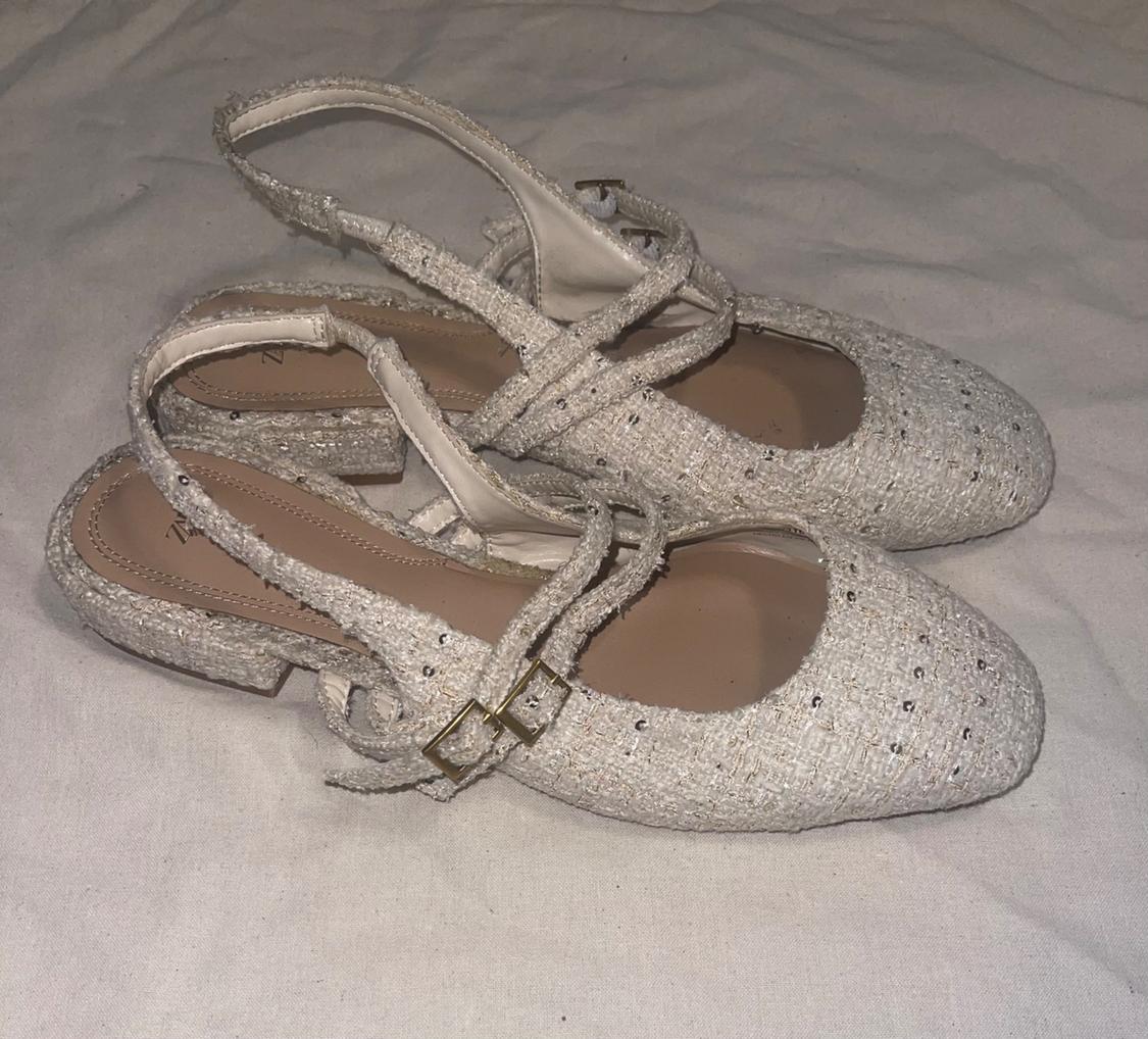 Cream-colored textured slingback flats with glitter and straps featuring gold buckles, resting on a light-colored wrinkled fabric.