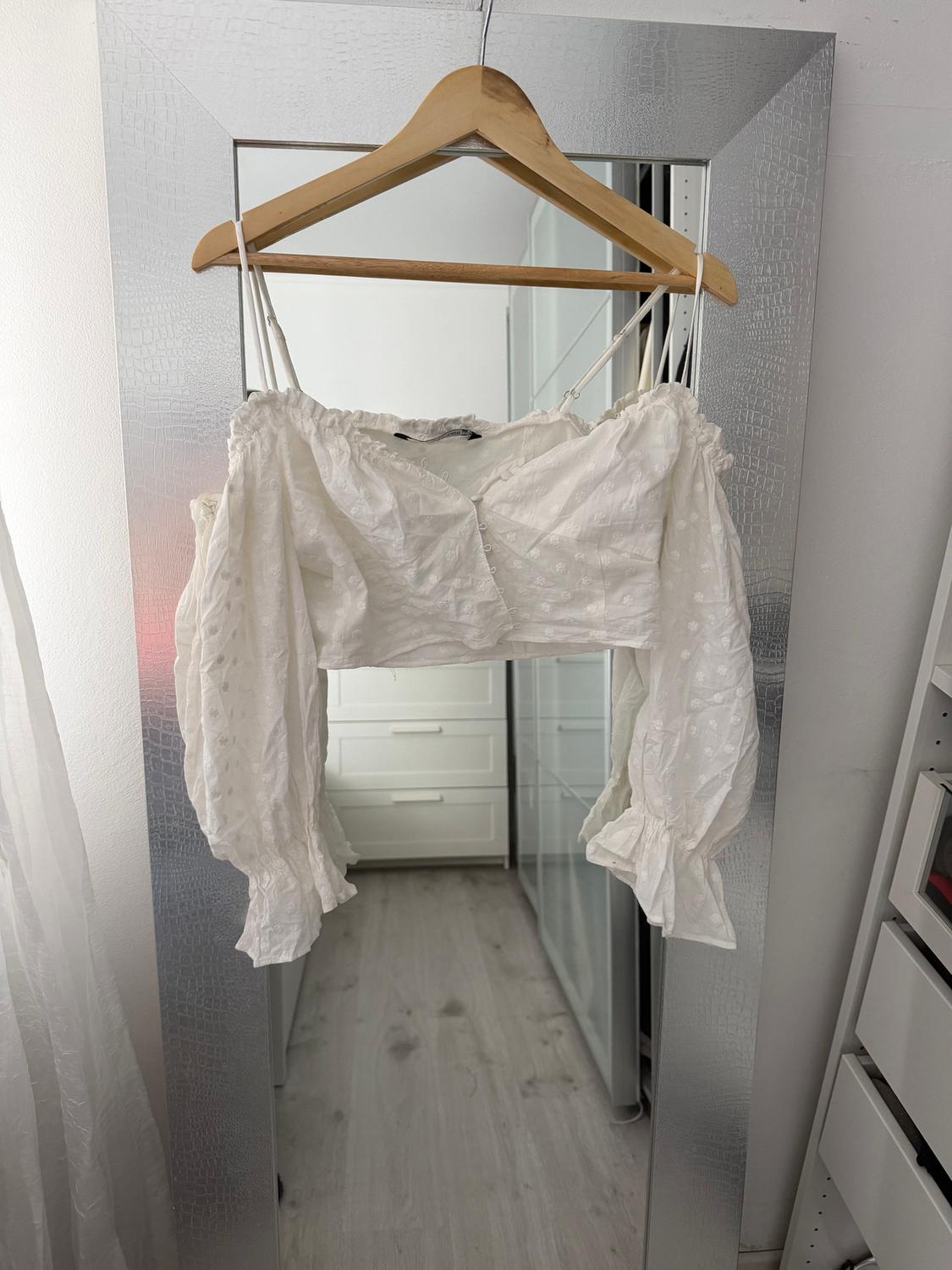 White blouse with long sleeves and lace details hanging on a wooden hanger in front of a mirror in a bright room.