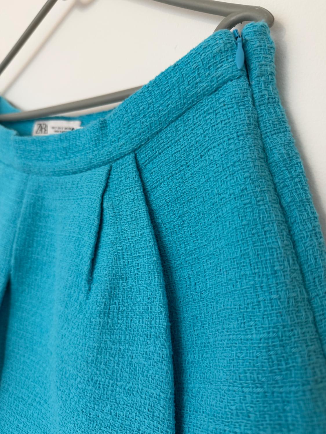 Close-up of a textured blue skirt with pleats and a side zip, hanging on a grey hanger against a white background. ZARA label visible.