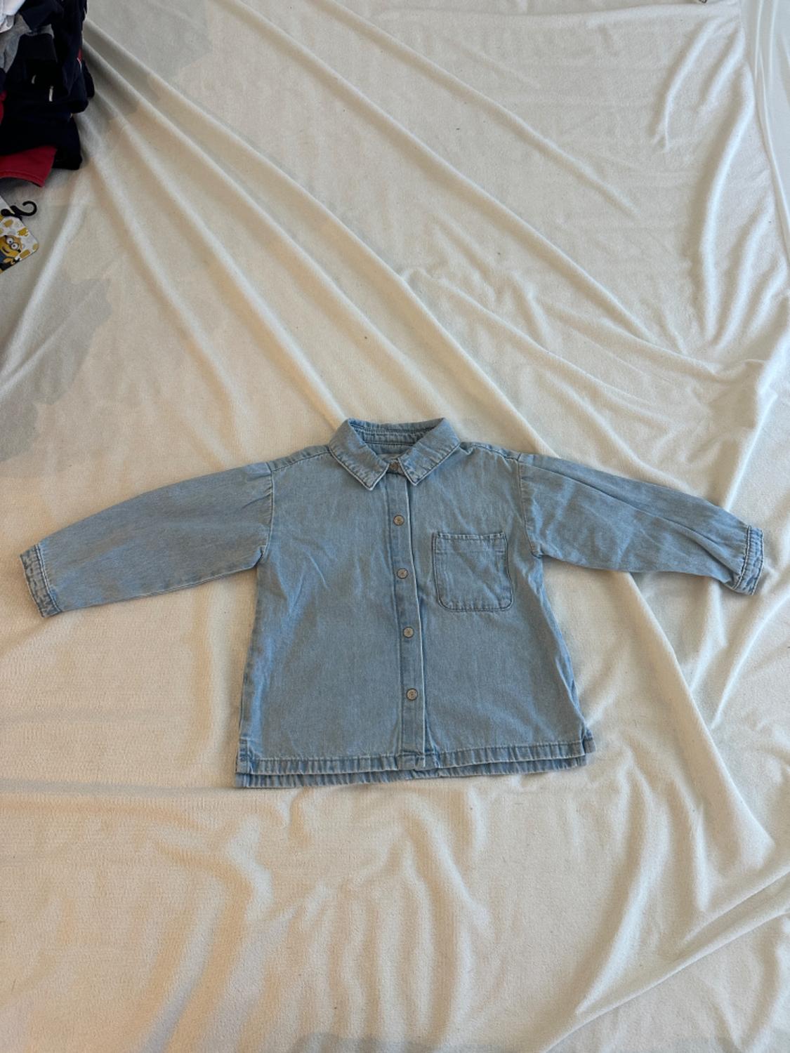 Light blue denim shirt for a boy with a front pocket, on wrinkled white fabric. Part of a stack of clothes visible on the left.