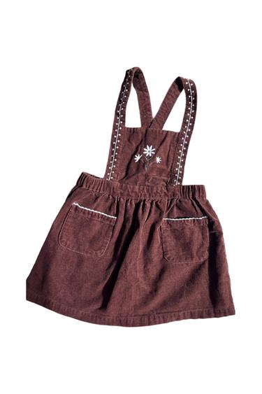 Brown cotton vest with crossed suspenders, embroidered white flowers on the chest, and two front pockets with white trim.
