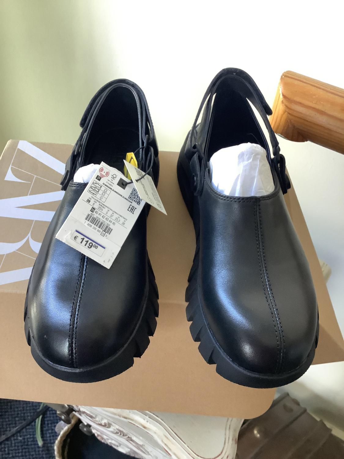 Black shoes with a serrated sole and a back strap on a box. They have paper stuffing and a label with the text EAC and €11,900.