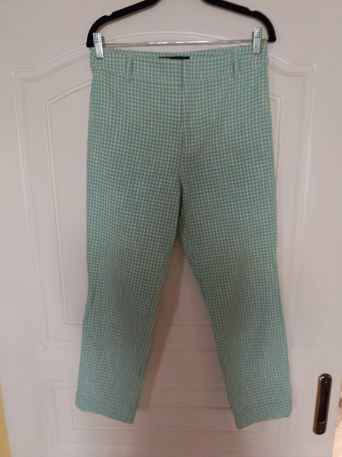 Green and white checked straight-leg trousers hanging on a black hanger in front of a white door.