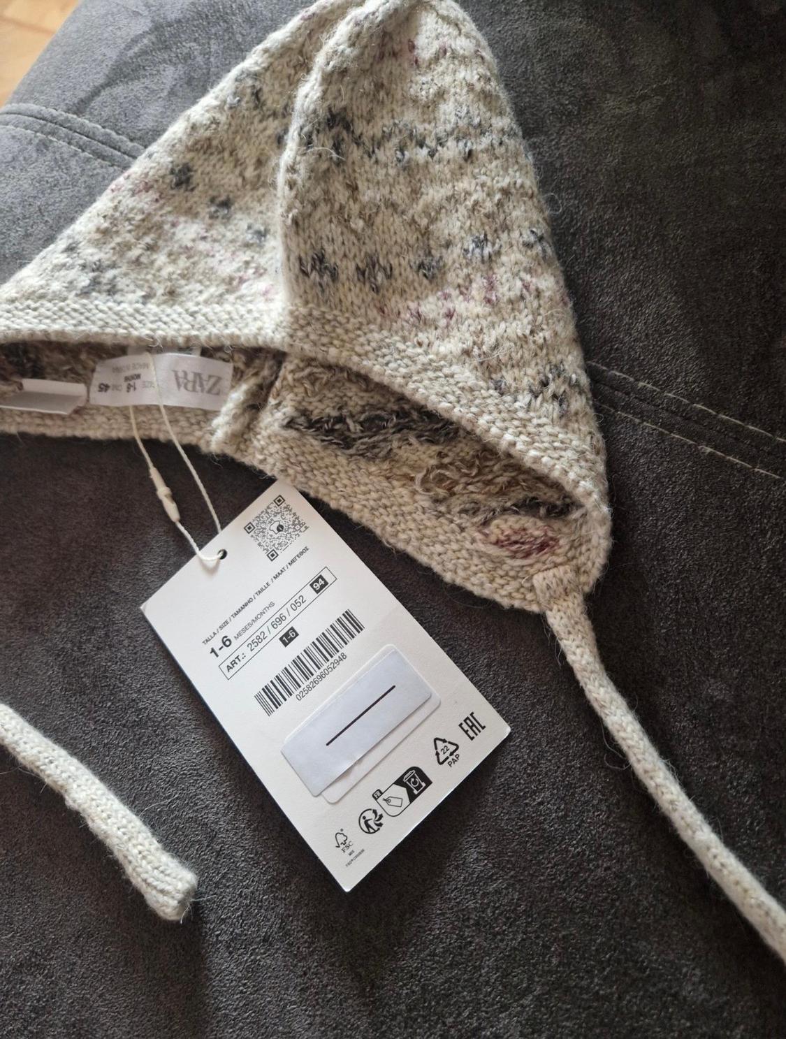 Knitted woolly hat in beige and grey tones on a dark grey sofa. Visible tag with barcode and Zara logo.