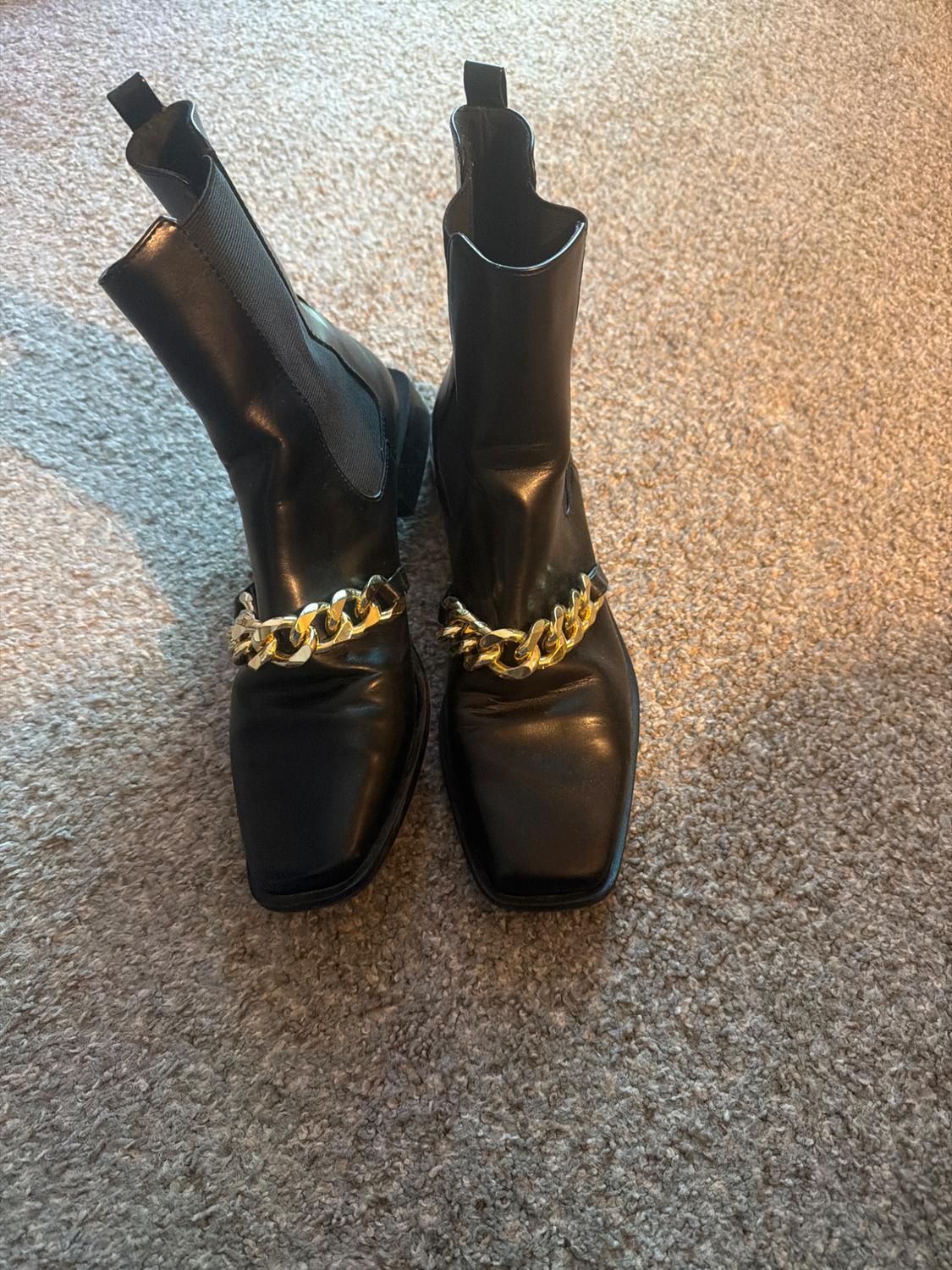 Black square-toe ankle boots with a gold chain on the instep and elastic panels, on a gray rug.
