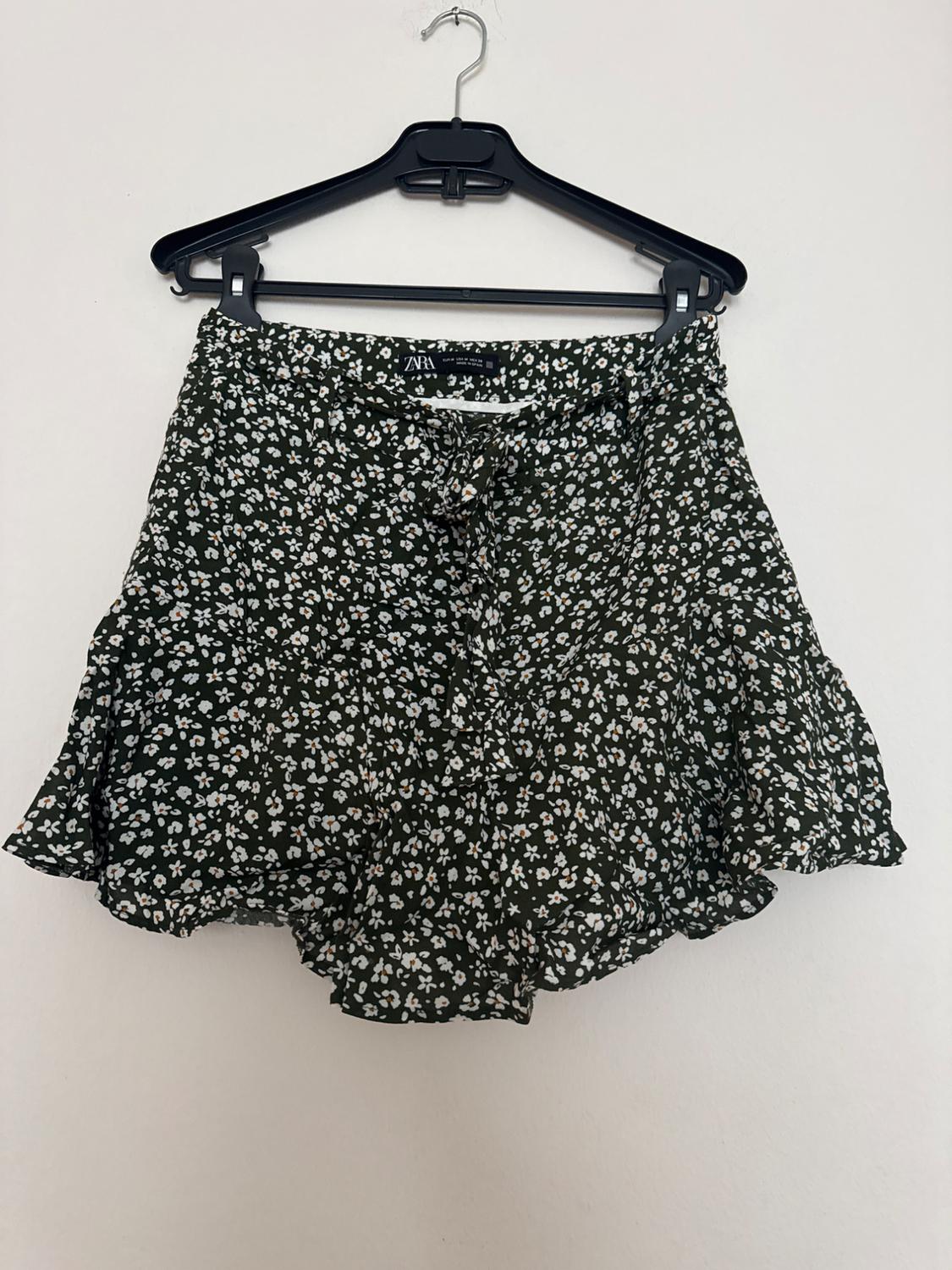 Short skirt with a white floral pattern on a dark green background, hanging on a black hanger against a white wall.