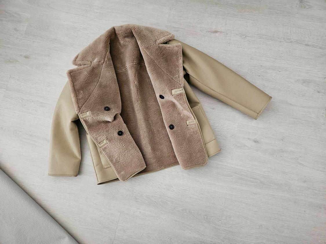 Beige jacket with wide lapels, soft faux-fur lining and dark buttons, open on a light wooden floor. Casual style.