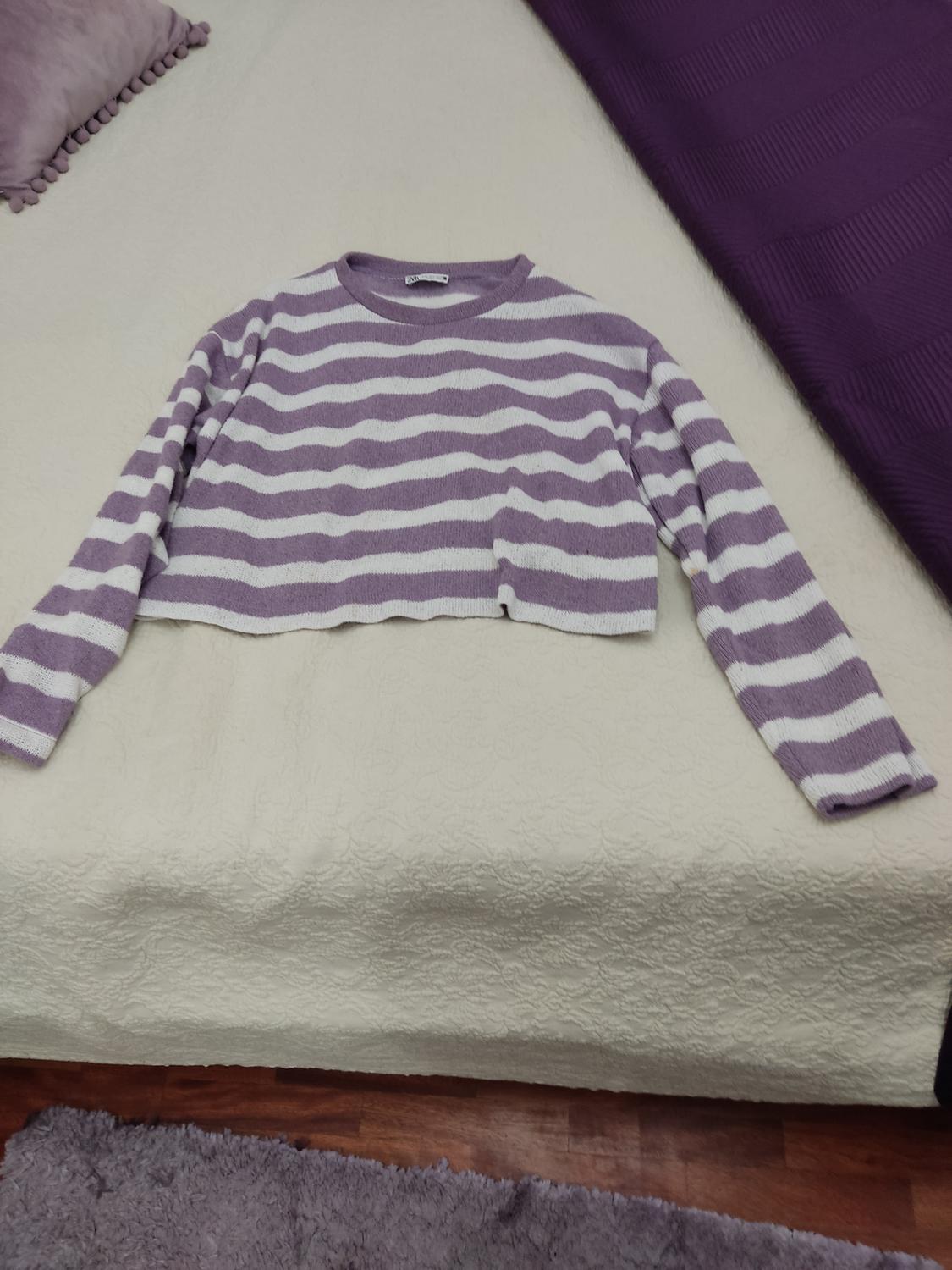 Purple and white striped jumper on a bed with a beige bedspread. Purple pillow and blanket in the background. Wooden floor.