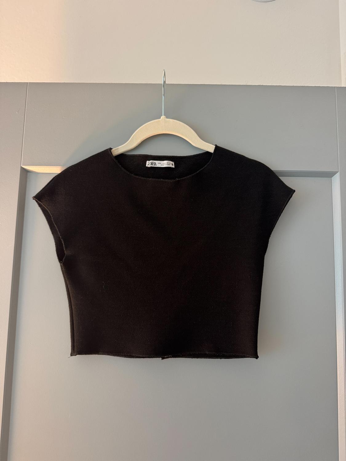 Black crop top hanging on a white hanger over a grey door. The label reads ZARA. Casual and minimalist style.