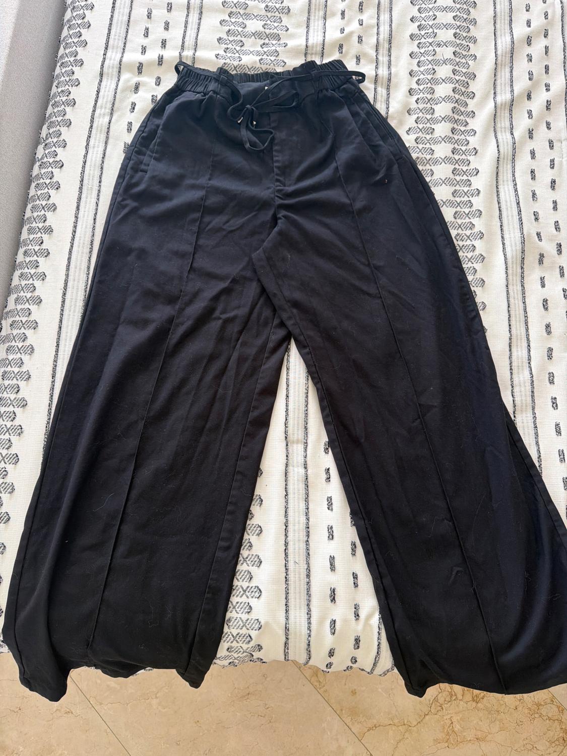 Wide-leg black trousers with an elasticated waistband, drawstring, and front seams, on a white blanket with black stripes in a casual setting.