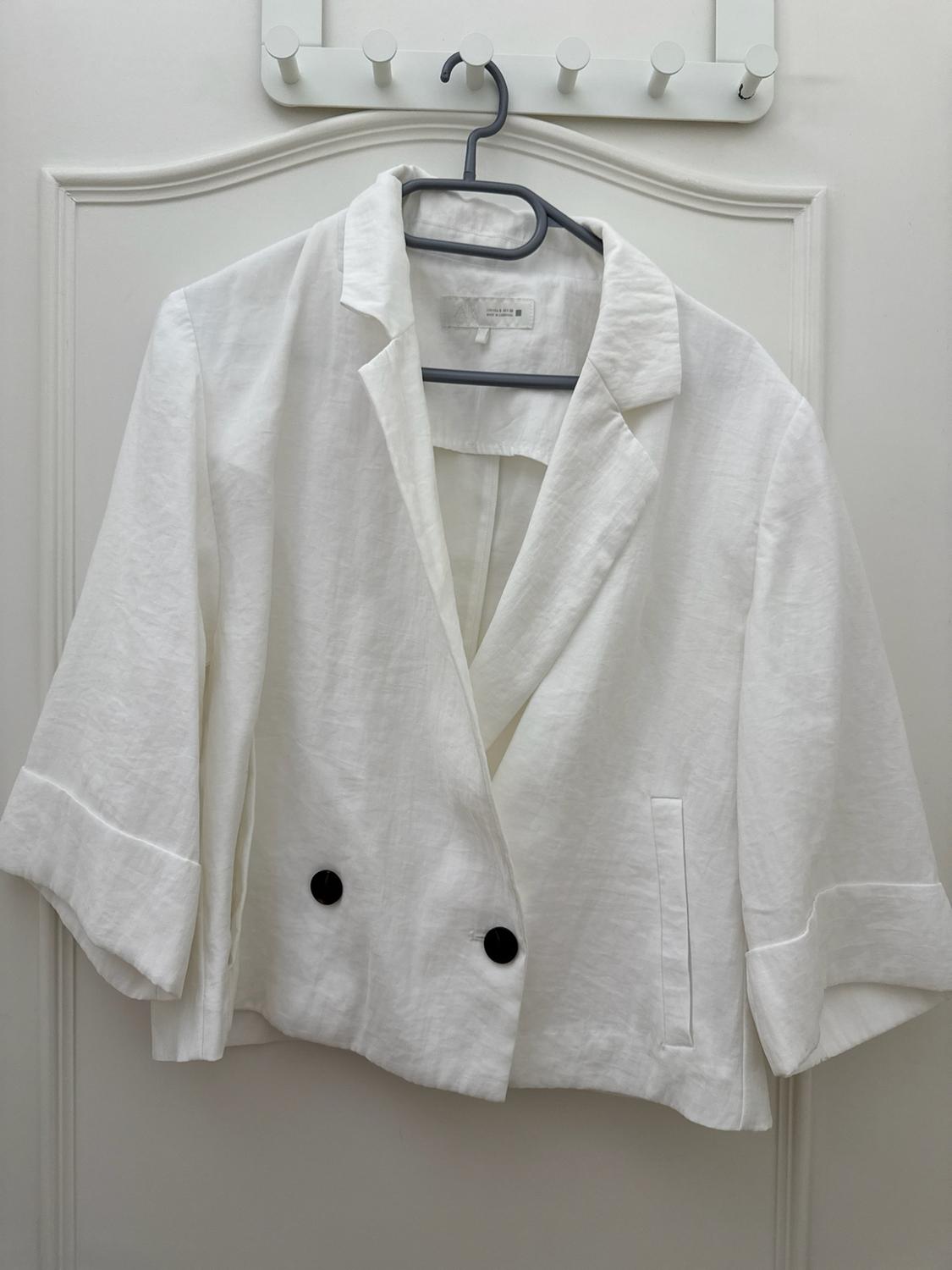 White jacket with lapels, three-quarter sleeves and two contrasting black buttons, hanging on a hanger over a white door.
