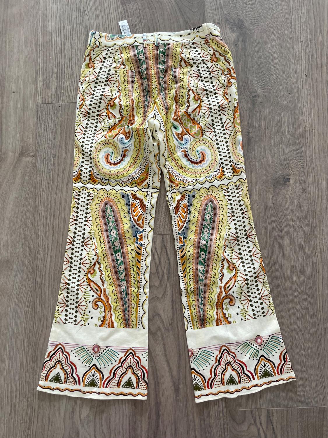 Printed pants with a paisley design in yellow, orange, and green hues on a white background, placed on a gray wooden floor.