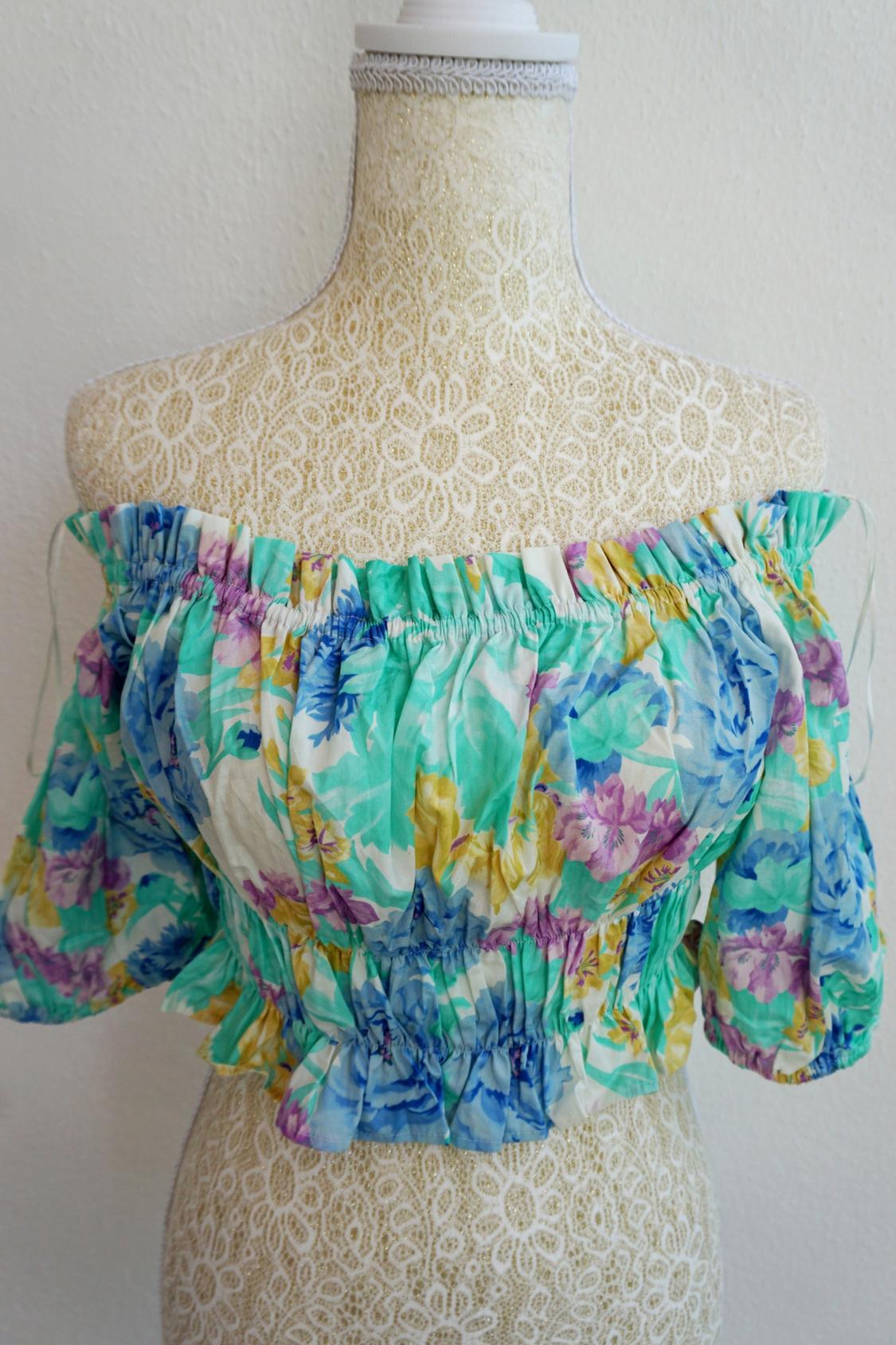 Off-the-shoulder blouse with a floral print in green, blue, and yellow tones on a beige lace mannequin.
