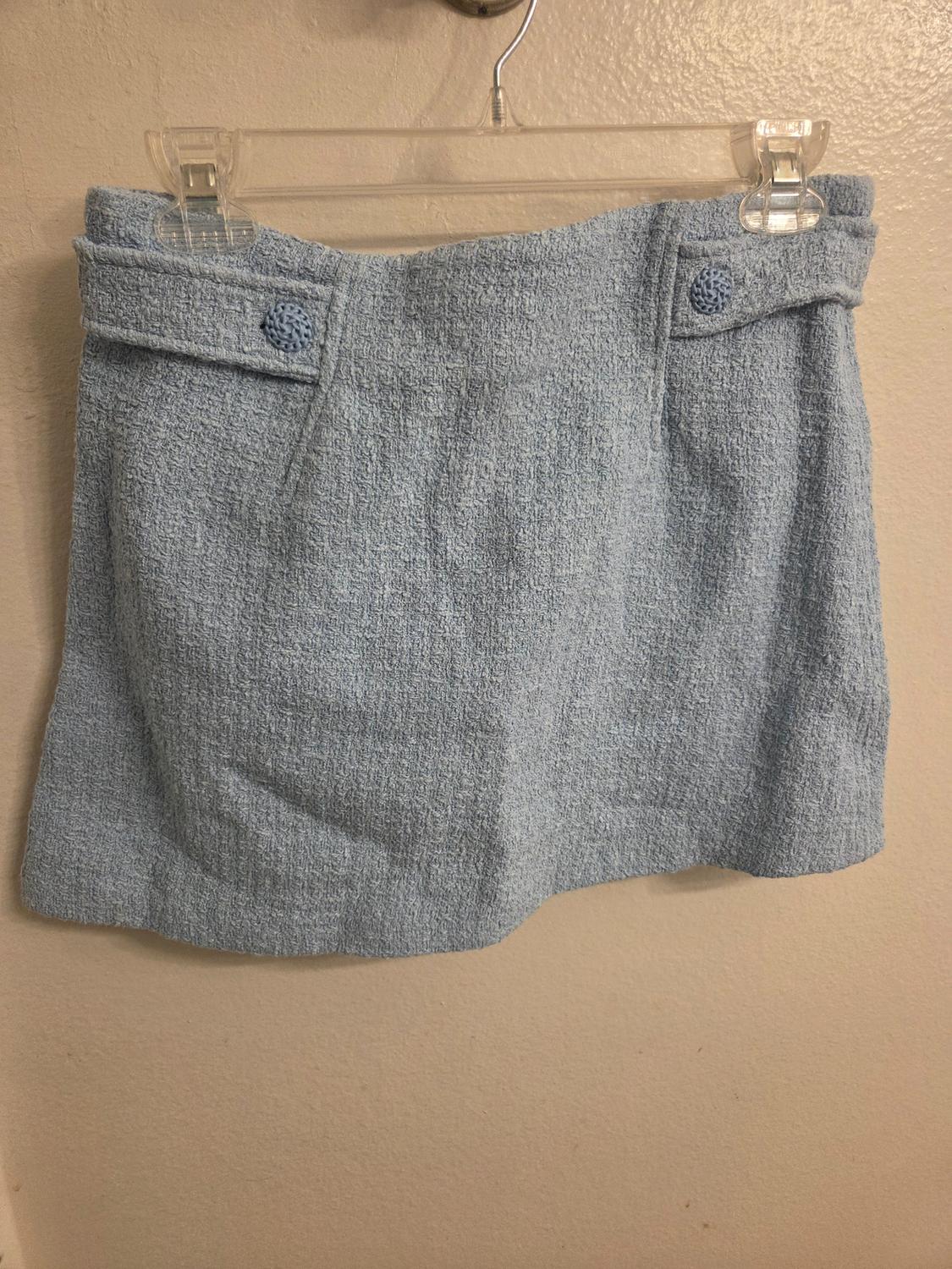 Light blue textured fabric short skirt hanging on a clear hanger against a beige wall.
