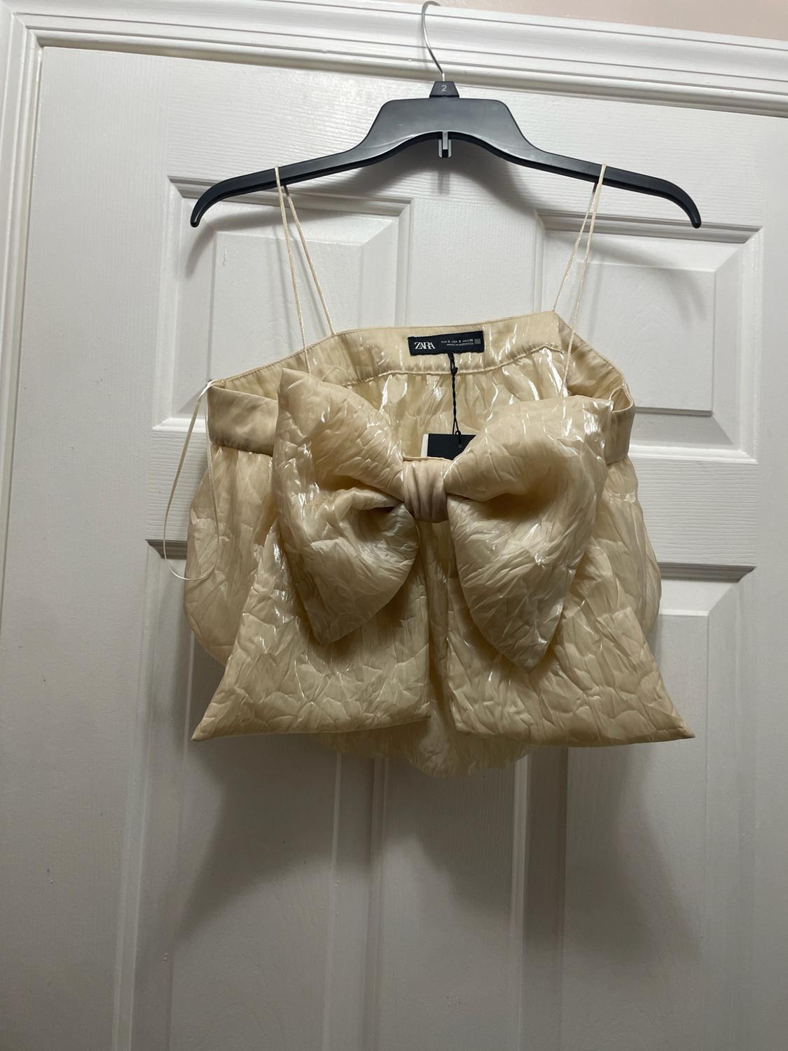 Shiny beige spaghetti-strap top with a large front bow and crinkled fabric, hanging on a hanger over a white door. ZARA label.