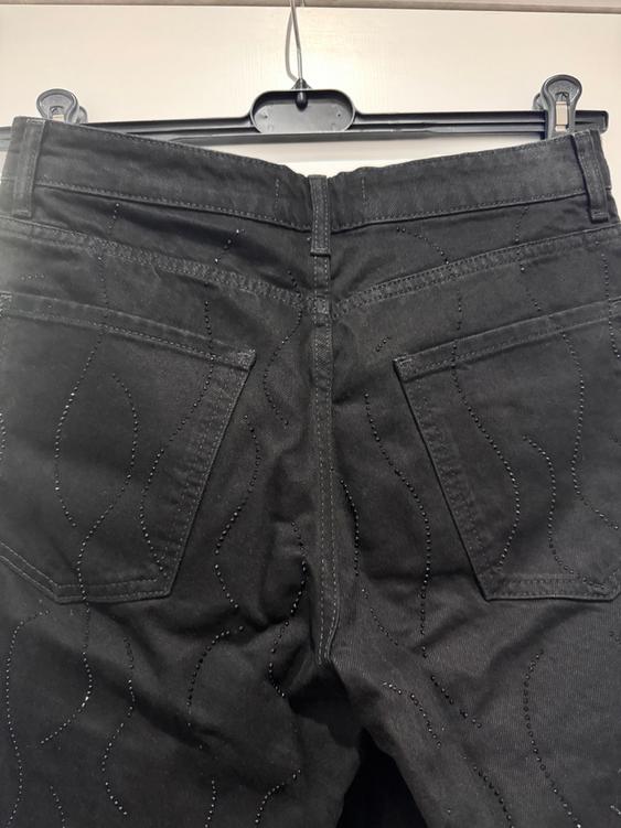 Black trousers hanging on a hanger, with wavy seam details and visible back pockets. White background.