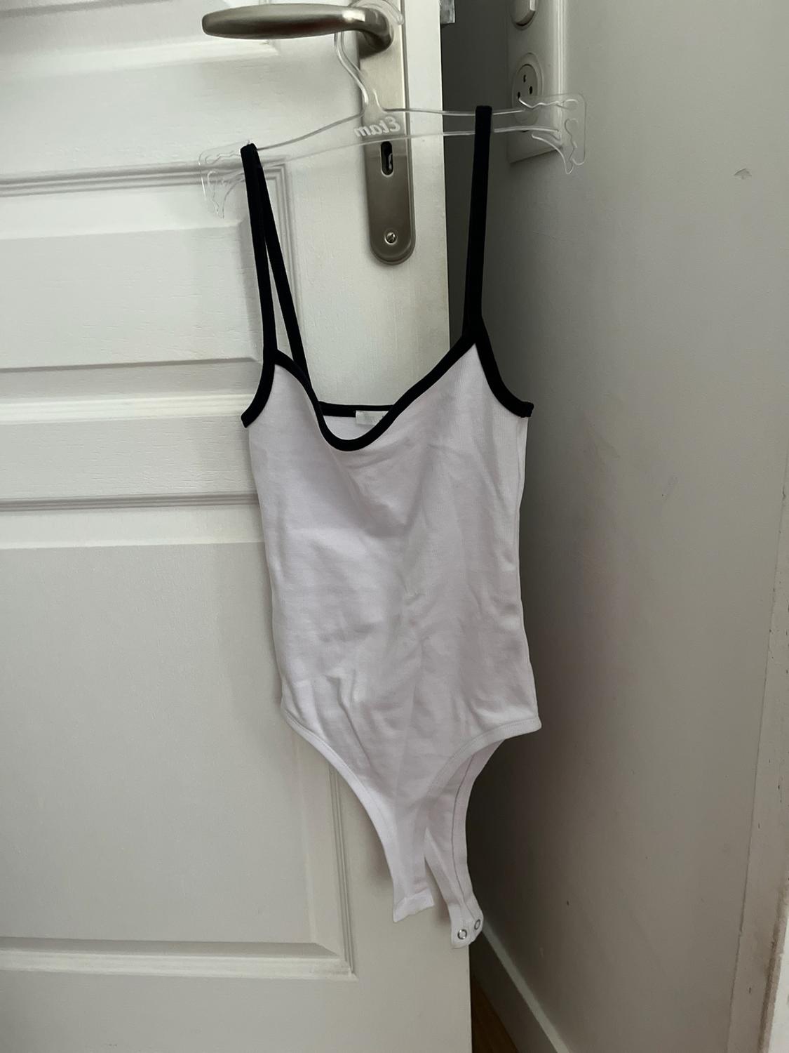 Casual white bodysuit with black trims and straps, on a clear hanger with "mo13" text, hanging from the silver handle of a white door.