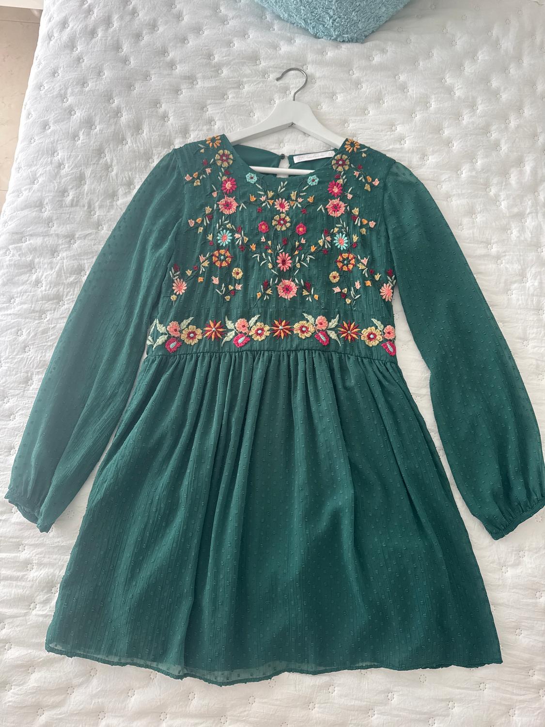 Green dress with colourful floral embroidery on the chest, long sleeves, on a white quilted bed.