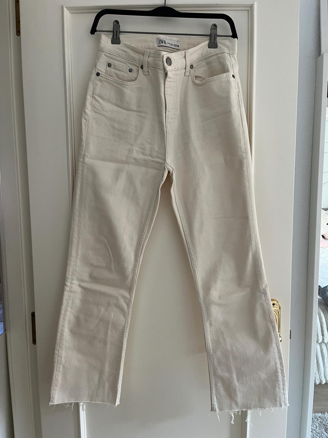 Beige denim trousers hanging on a black hanger over a white door. Visible label with the text "ZARA".