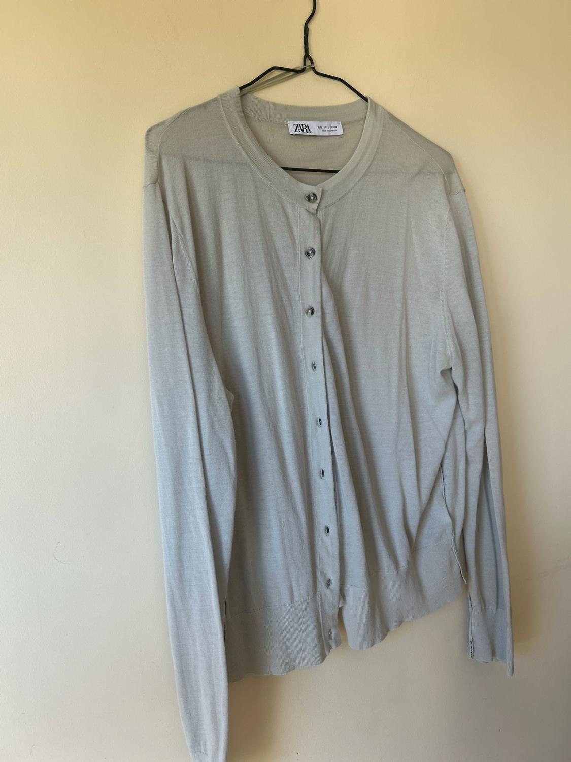 Lightweight beige cardigan with front buttons hanging on a wire hanger against a light-coloured wall. The label reads ZARA.