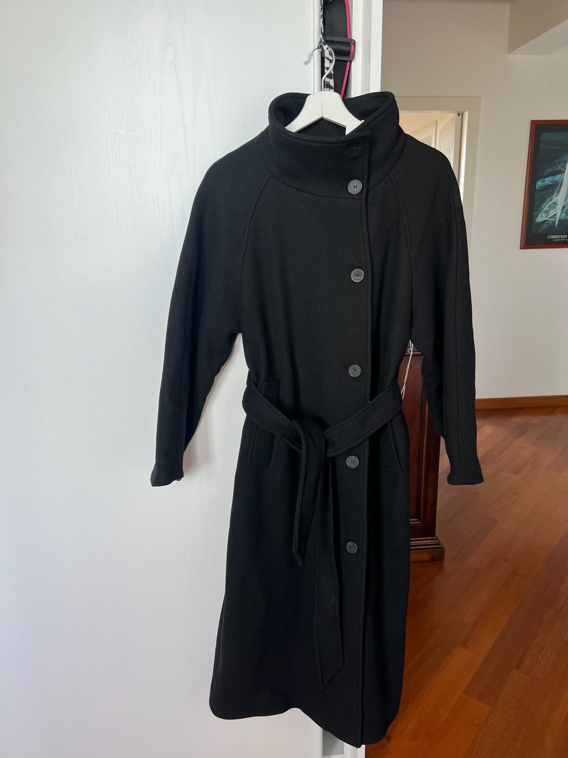 Long black coat with buttons and a belt, hanging on a white door. In the background, a wooden floor and a picture with the text CHRISTIAN.