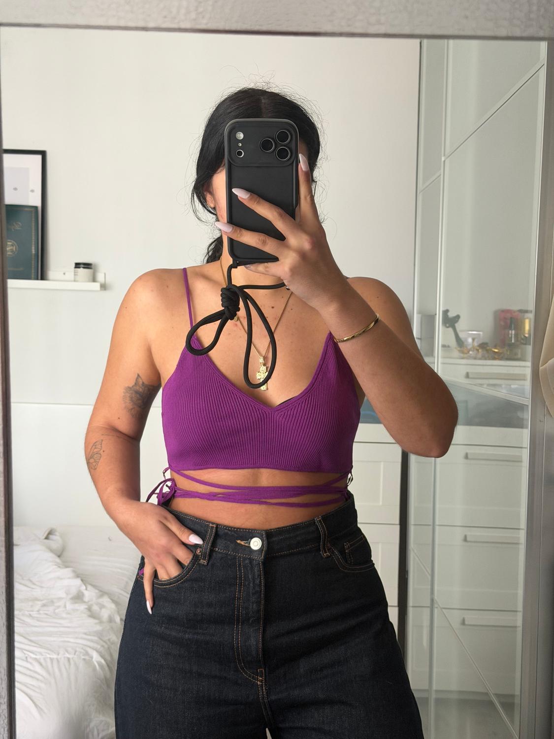 Woman in a purple top and dark jeans, taking a photo in her bedroom mirror. She's wearing a gold necklace and bracelet.