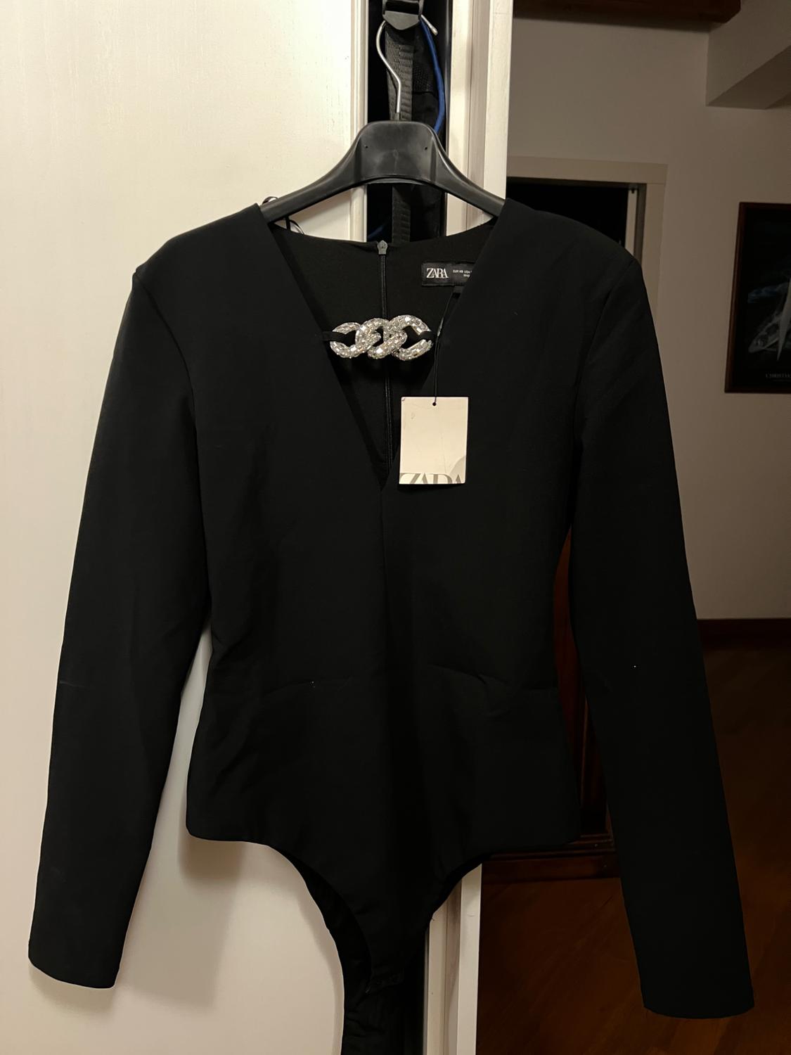 Black long-sleeved bodysuit on a hanger, with a silver brooch detail on the neckline. Visible label with the text "ZARA". Home interior background.