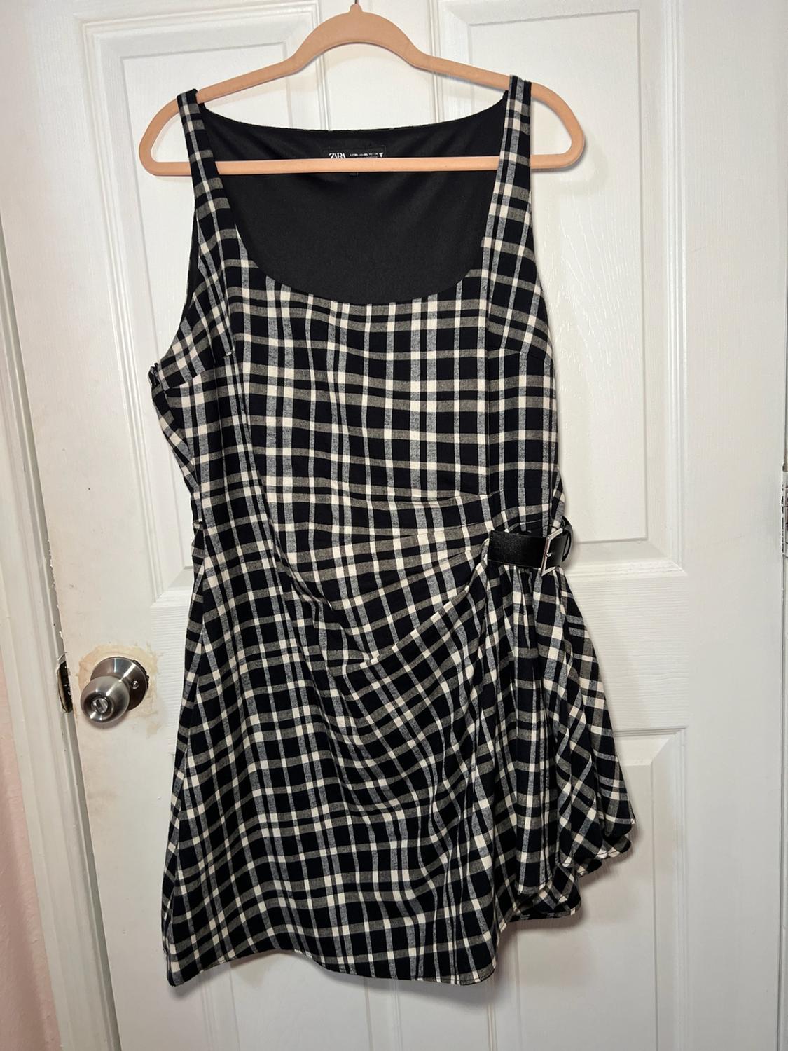 Black and white checked sleeveless dress with draping and a side buckle, hanging on a hanger on a white door. Visible text: ZARA.