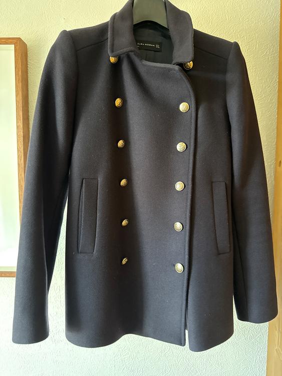 Black double-breasted coat with gold buttons hanging on a hanger. Visible label: "Zara Woman". Light wall background.
