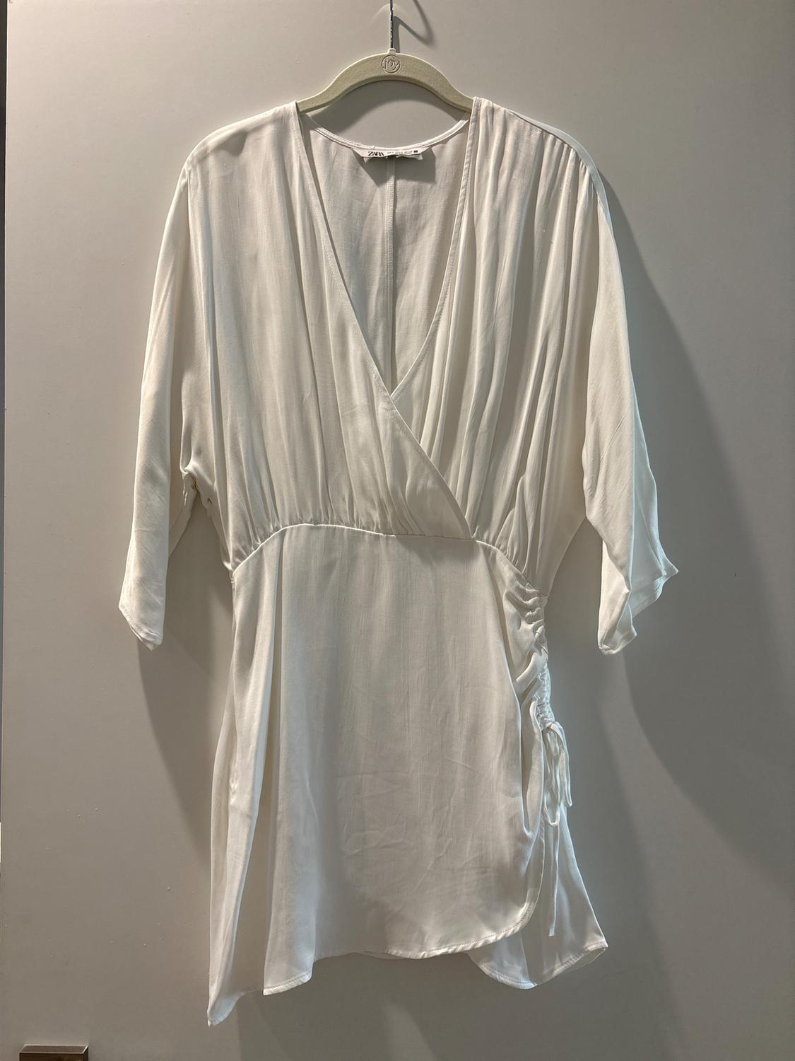 White three-quarter sleeve blouse with a V-neck and side ruching detail, hanging on a hanger against a gray background.
