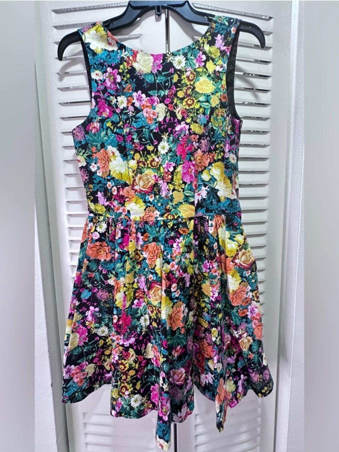 Sleeveless dress with a colourful floral print on a black background, hanging on a hanger in front of a white door.