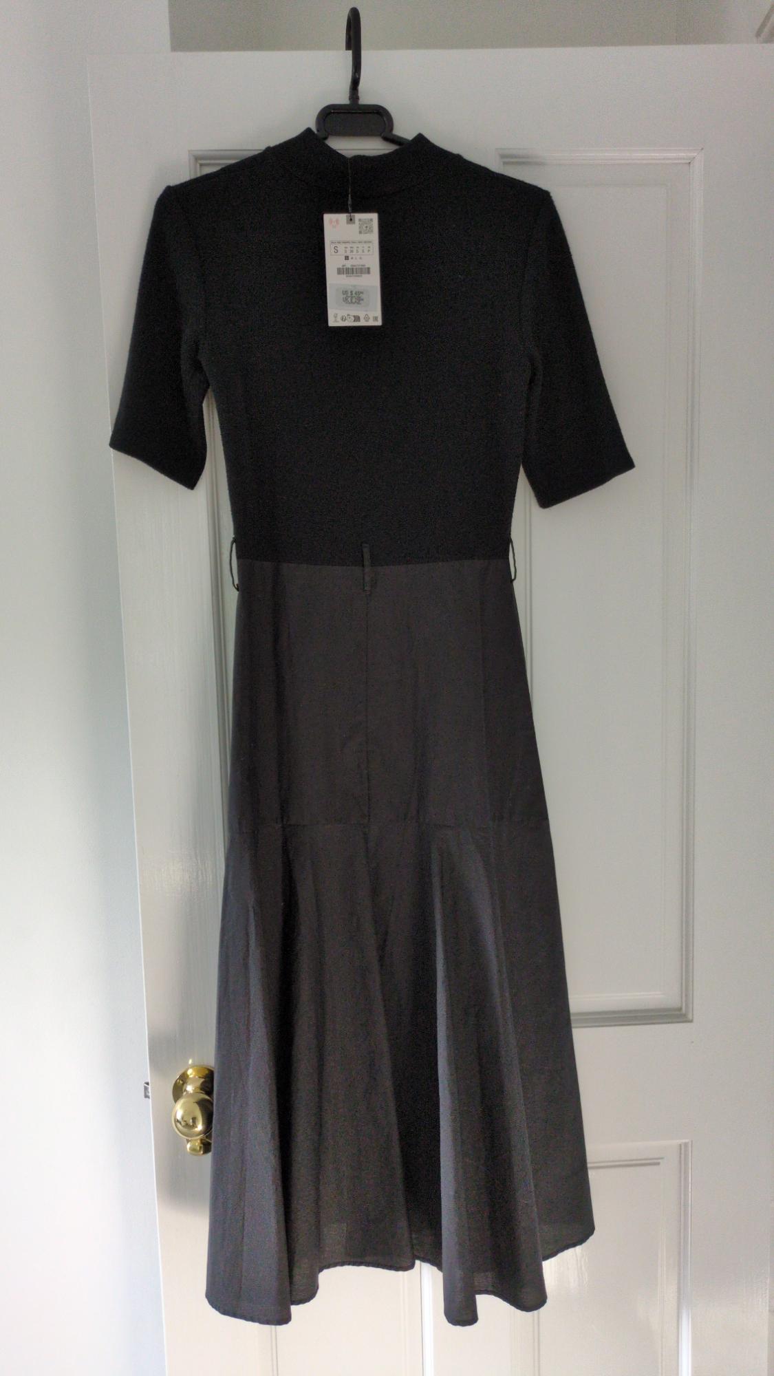 Black short-sleeved, high-neck dress with a label, hanging on a white door. It has a fitted bodice and a flared skirt.